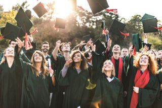 Large group of happy college students celebrating their graduation day outdoors while throwing their caps up in the air.